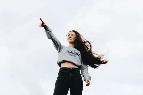 A person with long hair stands outdoors under a cloudy sky, wearing glasses and a Toronto sweatshirt. They are pointing forward, with a confident expression.