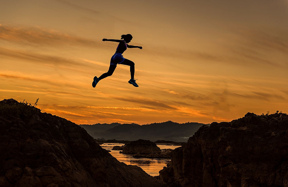 Silhouette of a person leaping across a rocky gap at sunset, with an orange sky and distant mountains in the background.