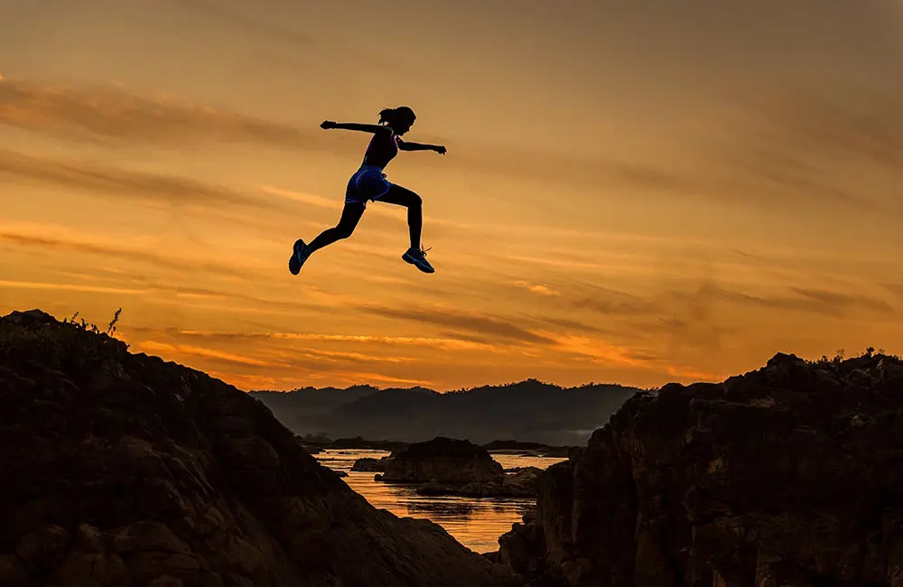 Silhouette of a person leaping across a rocky gap at sunset, with an orange sky and distant mountains in the background.